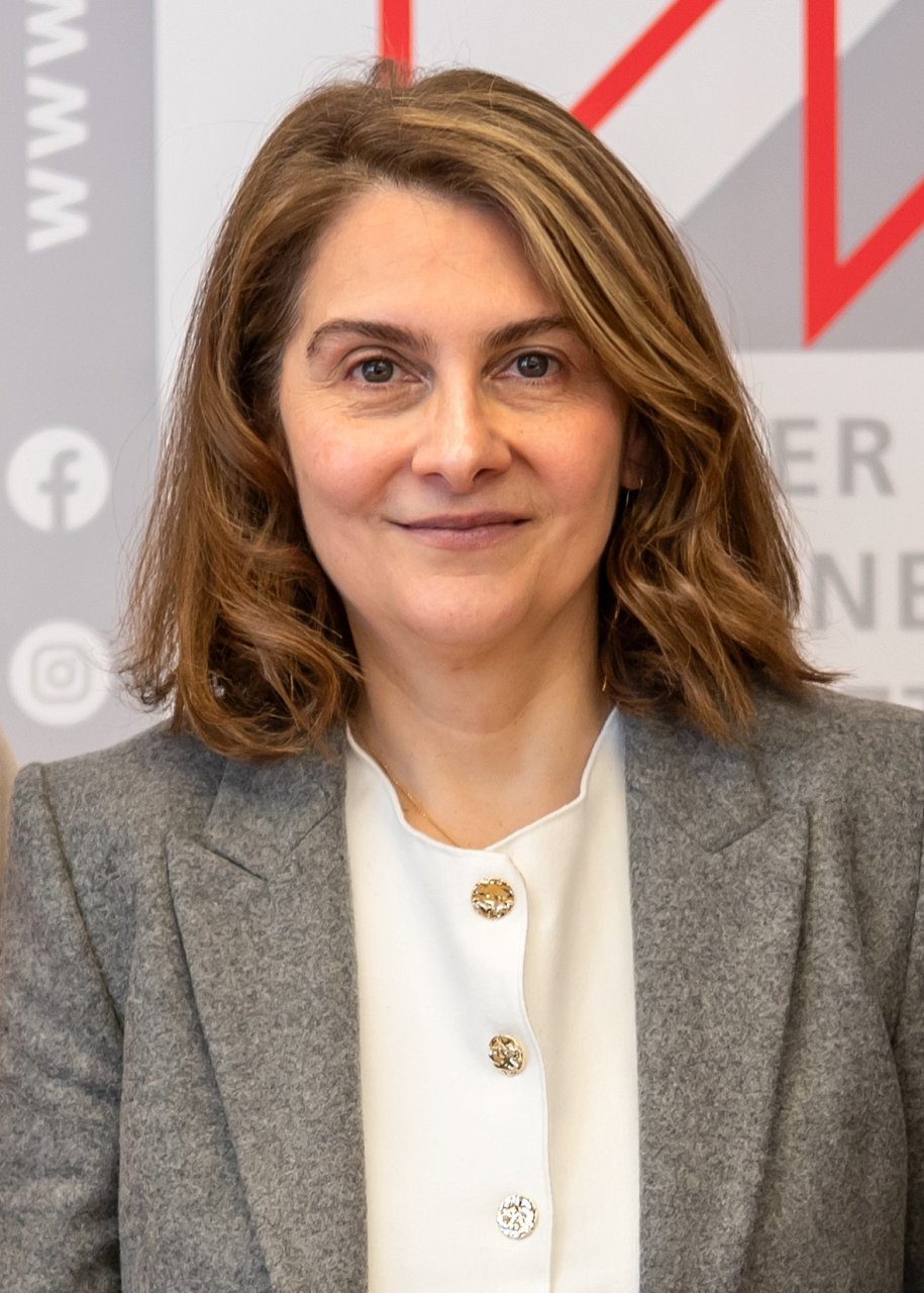 Referentin Katayoun Tonninger-Bahadori blickt direkt in die Kamera und im Hintergrund ist ein Plakat der Kammer für Ärztinnen und Ärzte in Wien zu sehen