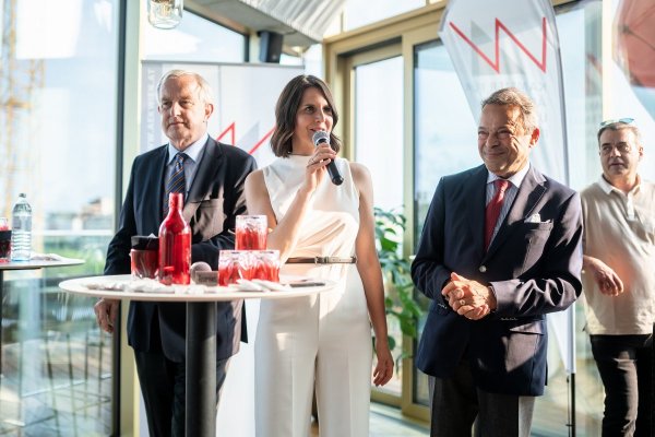 Gruppe von drei Personen, in der Bildmitte steht eine Sprecherin mit Mikrofon an einem Stehtisch.