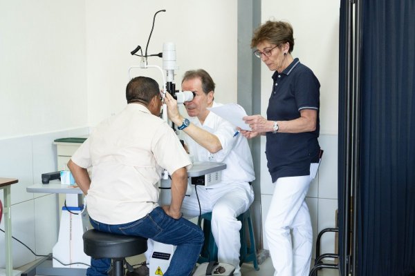 Drei Personen auf dem Foto - die vorderste Person wird gerade am Auge untersucht. Montserrat Masats steht gleich daneben und wirkt dabei mit.