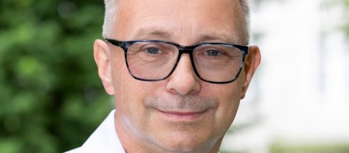 Ein Porträt des Arbeits- und Schmerzmediziners Richard Crevenna. Aufgrund des Hintergrunds kann geschlossen werden, dass die Aufnahme im Außenbereich stattgefunden hat - verschwommen ist ein Baum oder eine Hecke im Hintergrund zu sehen. 