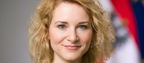 Die Bundesministerin Eva-Maria Holzleitner blickt in die Kamera, im Hintergrund sieht man die Österreichische Fahne, sowie einen kleinen Teil der EU-Flagge