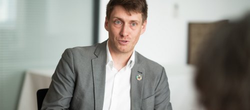 Mann mit Hemd und Sakko an einem Tisch sitzend, zum Interviewpartner sprechend.