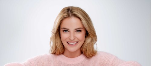 Die Ärztin und TV-Moderatorin Christine Reiler stützt sich mit beiden Armen auf einen Tisch auf und blickt lächelnd in die Kamera. Sie hat einen rosa, langärmeliges Oberteil an.