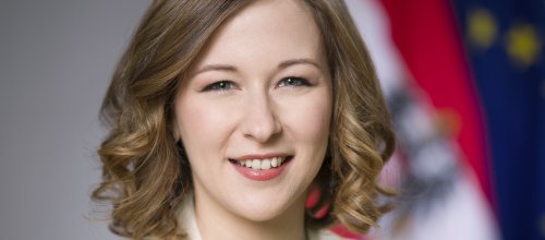 Frau mit Sakko vor österreichischer Fahne und EU-Fahne lächelt in die Kamera.