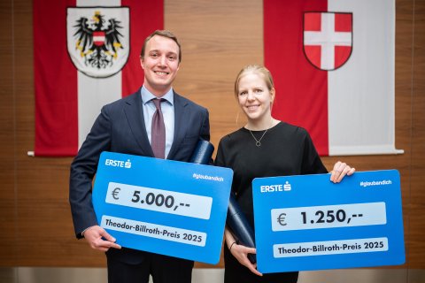 Die Preisträger*innen Moritz Muckenhuber und Anna Plötzl halten ihren Siegerscheck in der Hand und lächeln in die Kamera. Im Hintergrund sind zwei Flaggen (Österreich und Wien) zu sehen, die an der Wand hängen.