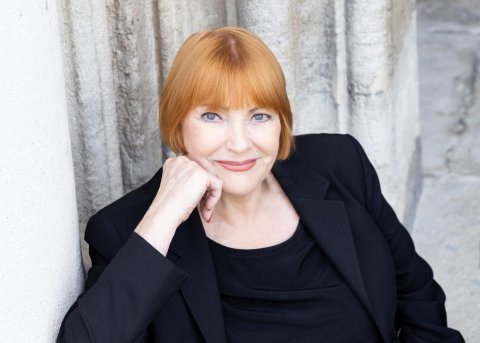 Auf dem Bild ist Chris Lohner zu sehen. Sie sitzt und blickt direkt in die Kamera. Ihre rechte Hand stützt ihre rechte Wange. Ihr Ausdruck wirkt freundlich und aufmerksam. Der Hintergrund ist neutral und lenkt nicht von der Person ab.