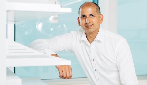 Der Mediziner Wali Malik sitzt in einem Labor, weiß bekleidet und blickt direkt in die Kamera.